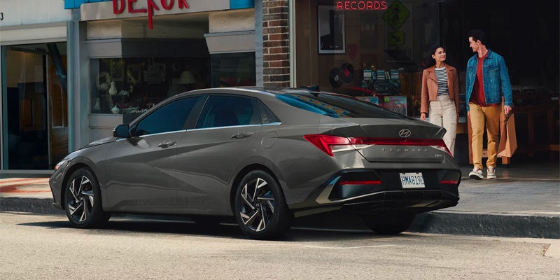 Hyundai Elantra Hybrid is parked curbside. Two people with shopping bags walk on the sidewalk in front of a 