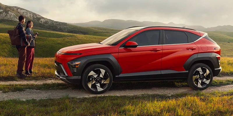 Hyundai Kona parked on a dirt road, facing right. A couple stands left of the car, admiring the green hills.