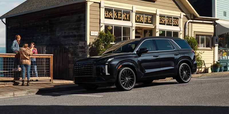 Hyundai Palisade is parked angled on a street before a 'BAKERY CAFE ESPRESSO' shop with light-colored siding. Three girls chat on a wooden deck left of the SUV, with a dark wooden building behind them.