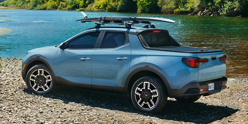 Hyundai Santa Cruz pickup truck with a surfboard on its roof rack, parked on a rocky shoreline next to blue water.
